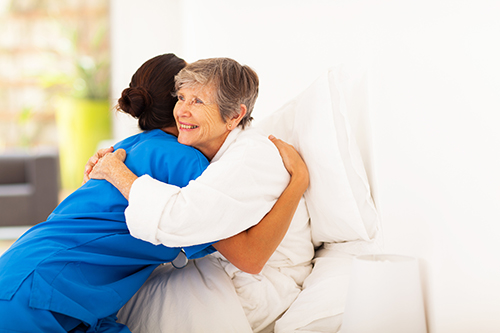 CNA in blue scrubs leaned in hugging an elderly woman wearing a white robe